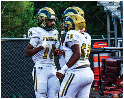 Three people wearing white and gold football uniforms and helmets are standing and interacting near a chain-link fence with trees and bleachers in the background. Description generated by AI