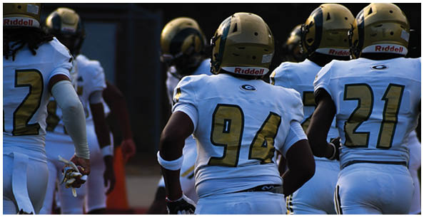 Four people wearing white football uniforms with gold helmets and large numbers on their backs are standing on a field. Description generated by AI