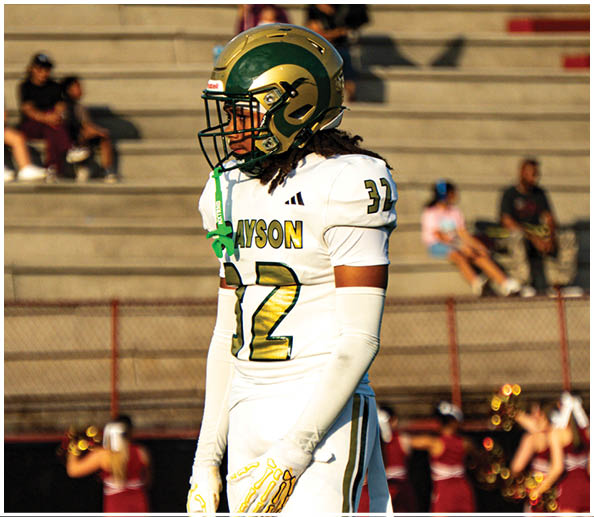 A person in a white football uniform with the number 12 and a gold helmet stands on a football field with blurred spectators and cheerleaders in the background. Description generated by AI