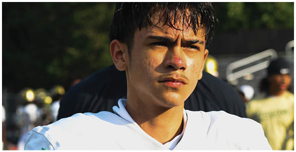 A person with short dark hair wearing a white shirt is looking intently ahead outdoors with blurred people and greenery in the background. Description generated by AI