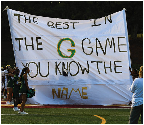 People in cheerleading uniforms hold a large white banner on a sports field that reads “THE BEST IN THE G GAME YOU KNOW THE NAME" with the letter G highlighted in green and gold. Description generated by AI