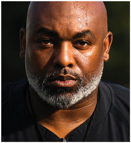 A person with a bald head and a gray beard is looking directly at the camera with a serious expression and visible sweat on their forehead. Description generated by AI