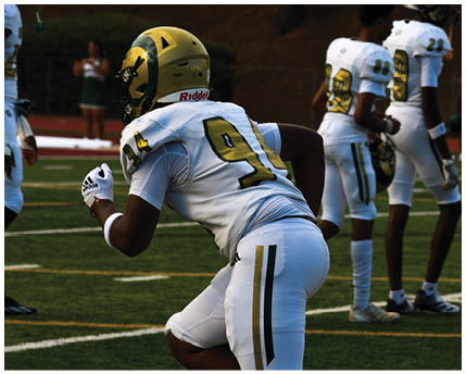 A person in a white and gold football uniform is crouched on a field with other players in similar uniforms standing in the background. Description generated by AI