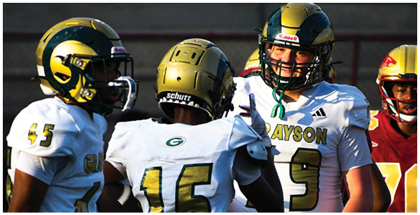 Four people wearing football helmets and uniforms are engaged in conversation on a field with bleachers in the background. Description generated by AI