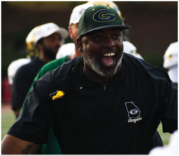 A person wearing a dark jacket and a green cap with a “G" logo is smiling widely and appears to be enthusiastically engaging with others on a sports field. Description generated by AI
