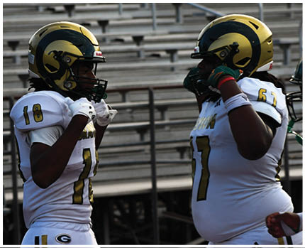 Two people wearing football helmets and white jerseys with gold and green accents are facing each other in a stadium with empty bleachers in the background. Description generated by AI