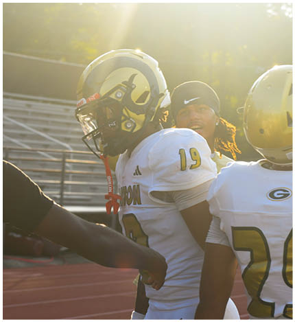 People in football uniforms are on a field with sunlight streaming through trees in the background. Description generated by AI