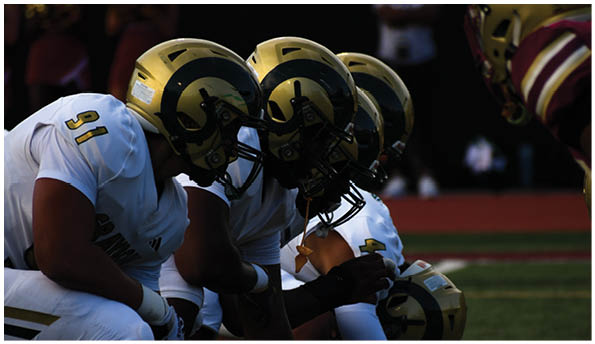 Four people wearing football helmets and uniforms are lined up facing an opponent on a field. Description generated by AI