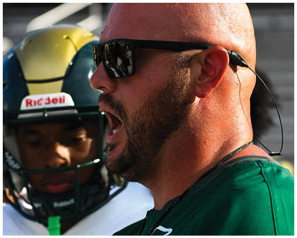 A person wearing sunglasses and a headset is speaking intensely while another person in a football helmet looks on in the background. Description generated by AI