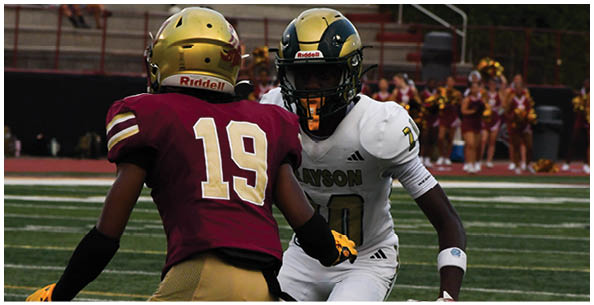 Two people wearing football helmets and uniforms face each other on a football field with cheerleaders and spectators in the background. Description generated by AI