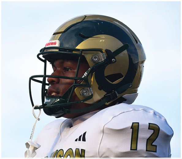 A person wearing a football helmet and white jersey with the number 12 stands against a clear sky. Description generated by AI