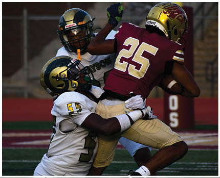 Two people in white and gold football uniforms tackle a person in a maroon and gold football uniform holding a football near a goalpost on a field. Description generated by AI