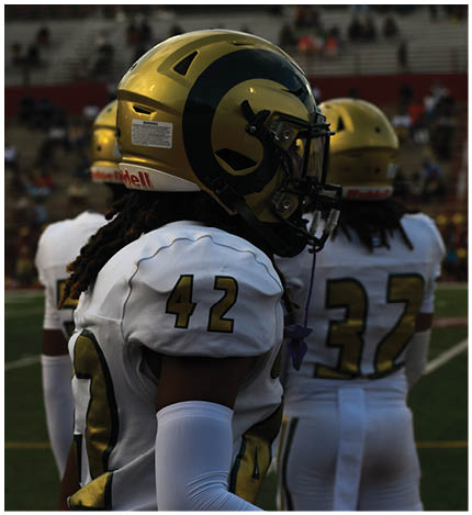 A person wearing a gold football helmet with a black ram horn design and a white jersey with the number 42 stands on a field with other players in the background. Description generated by AI