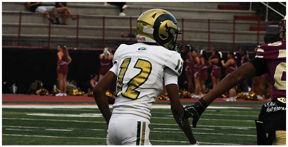 A person wearing a white football uniform with the number 12 and a gold helmet is on a field with cheerleaders and people sitting in bleachers in the background. Description generated by AI