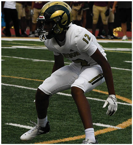 A person wearing a white football uniform with the number 12 is crouched in an athletic stance on a green turf field marked with white and yellow lines. Description generated by AI