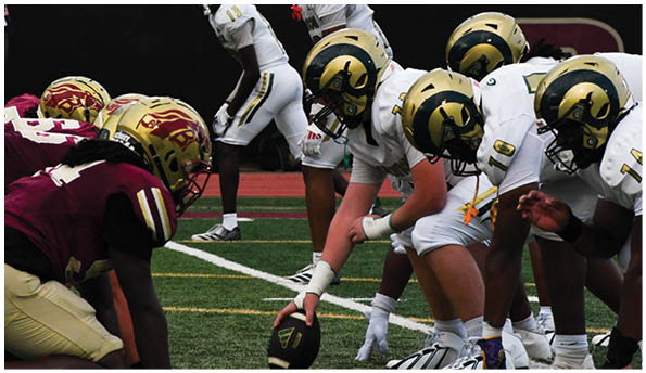 Two opposing American football teams are lined up facing each other on a field, with one team wearing white uniforms and gold helmets and the other team wearing maroon uniforms and gold helmets. Description generated by AI