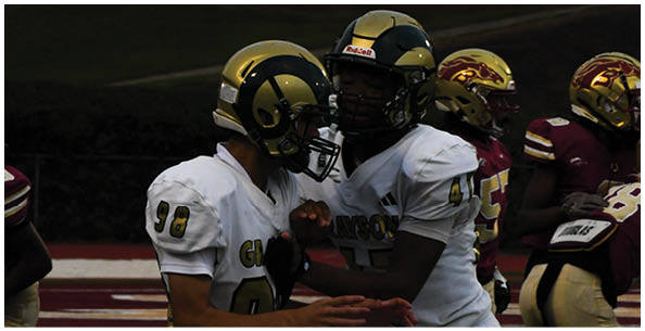 People wearing football helmets and uniforms are gathered closely on a field with a dark background. Description generated by AI
