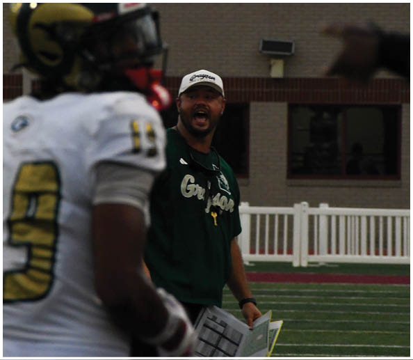 A person wearing a green sweatshirt and white cap is holding papers and appears to be speaking on a sports field while a player in a white and gold football uniform stands nearby. Description generated by AI