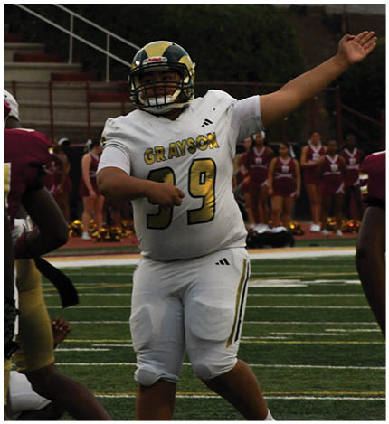 A person in a white football uniform with the name Grayson and number 39 is raising one arm and pointing with the other on a football field while other players and cheerleaders stand in the background. Description generated by AI
