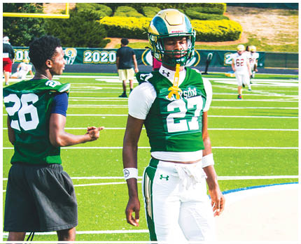 Two people wearing green football jerseys stand on a football field with one person wearing a helmet and full gear while the other wears shorts and gestures with their hand. Description generated by AI