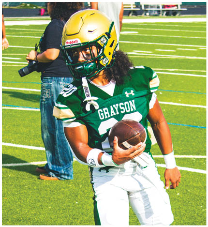 A person wearing a green and white football uniform with the name Grayson is holding a football and standing on a field with another person in the background holding a camera. Description generated by AI