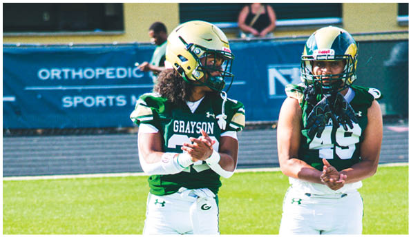 Two people in football uniforms with helmets stand on a field clapping, one wearing a green jersey with “GRAYSON" and the other wearing a black jersey with the number 49. Description generated by AI