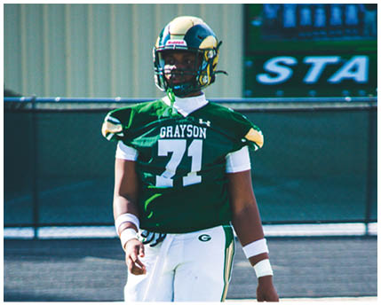 A person wearing a green football jersey with the number 71 and the word Grayson stands on a field with a helmet and white pants. Description generated by AI
