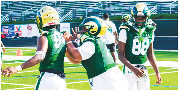 Three people wearing green football uniforms and helmets are on a football field with empty bleachers in the background. Description generated by AI
