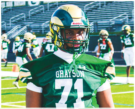 A person wearing a green football jersey with the number 71 and a helmet stands on a football field with other players in the background. Description generated by AI