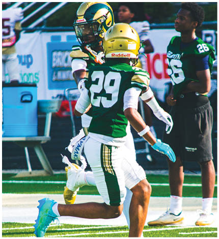 An adult wearing a green and white football uniform with the number 29 is running on a football field while two other adults stand nearby. Description generated by AI
