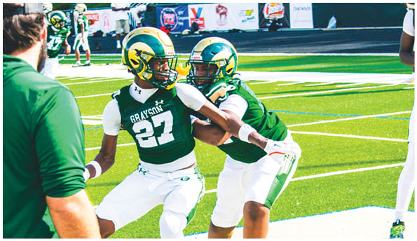 Two people in green and white football uniforms with helmets are on a football field, with one person appearing to hold or block the other while a person in a green shirt watches nearby. Description generated by AI
