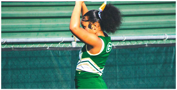 A person wearing a green and white cheerleading uniform is raising their arms, standing in front of a chain-link fence. Description generated by AI