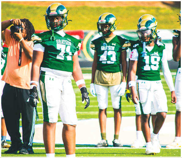 Three people wearing green football jerseys and white pants stand on a football field with a person in casual clothing nearby. Description generated by AI