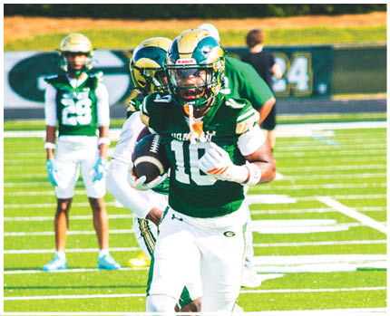 A person wearing a green and white football uniform is running on a field while holding a football, with another person closely behind. Description generated by AI