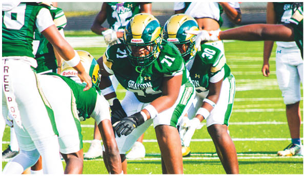 Several people in green and white football uniforms are positioned on a grassy field, preparing for a play. Description generated by AI