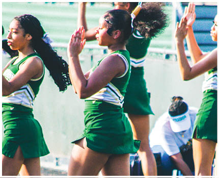 People in green and white cheerleading uniforms are performing outdoors while another person in casual clothing is seated nearby. Description generated by AI