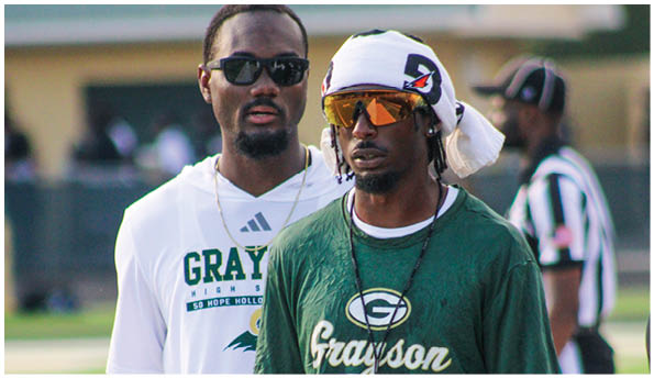 Two adults wearing sports apparel and sunglasses stand outdoors on a field with a referee visible in the background. Description generated by AI