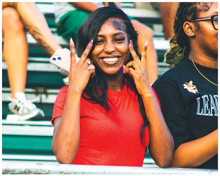 A person wearing a red shirt and jeans is smiling and making a peace sign with both hands while standing behind a metal fence in a stadium setting. Description generated by AI