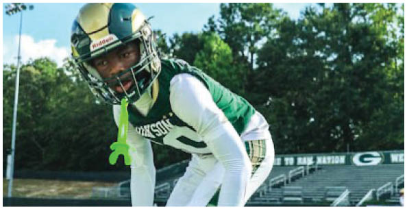 A person wearing a green and white football uniform is crouched on a field with empty bleachers and trees in the background. Description generated by AI