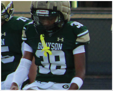 A person wearing a green football jersey with the number 38 and the word Grayson is sitting with a white arm sleeve and a yellow mouthguard hanging from the helmet. Description generated by AI