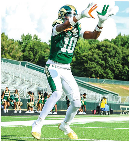 A person wearing a green and white football uniform with the number 10 is jumping with arms extended to catch a football on a field with cheerleaders and empty bleachers in the background. Description generated by AI
