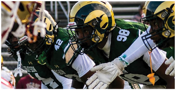 Four people in football uniforms and helmets are lined up in a ready stance on a field, preparing for a play. Description generated by AI