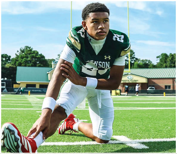 A person in a green and white football uniform is crouched on a football field with a goalpost and building in the background. Description generated by AI