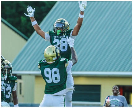 Two people in football uniforms are jumping to catch a ball on a field while another person stands nearby holding a drink and smiling. Description generated by AI