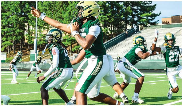 Several people in green and white football uniforms are actively engaged in a practice or game on a sunny outdoor field with bleachers in the background. Description generated by AI