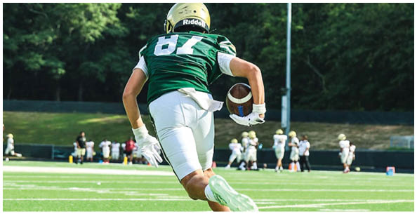 A person wearing a green sports jersey and helmet is running on a grassy field while holding a football. Description generated by AI