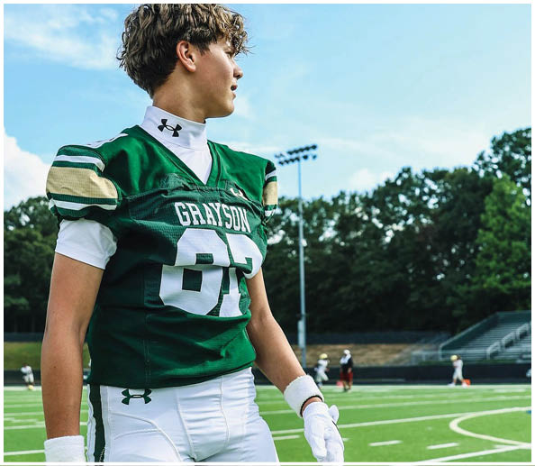 A person wearing a green football jersey with the name Grayson and the number 87 is standing on a football field with other players in the background. Description generated by AI