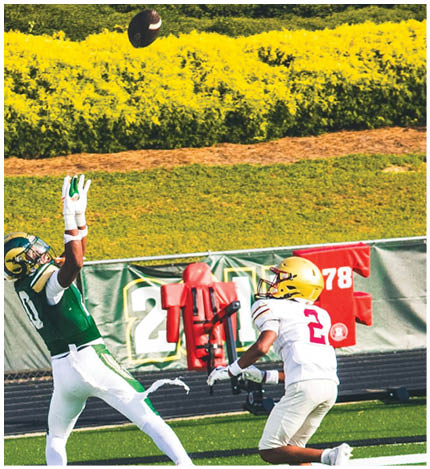 A person wearing a white football uniform with the number 2 and a gold helmet is running on a green field while another person in a green uniform is reaching out with their arm extended. Description generated by AI