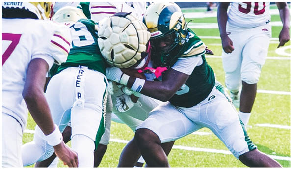 People in football uniforms are engaged in a close physical play on a grassy field with one player holding the ball tightly. Description generated by AI