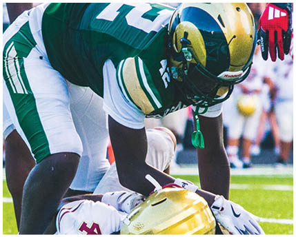 A person wearing a green and white football uniform with a gold helmet is bent forward on a field, preparing for a play. Description generated by AI